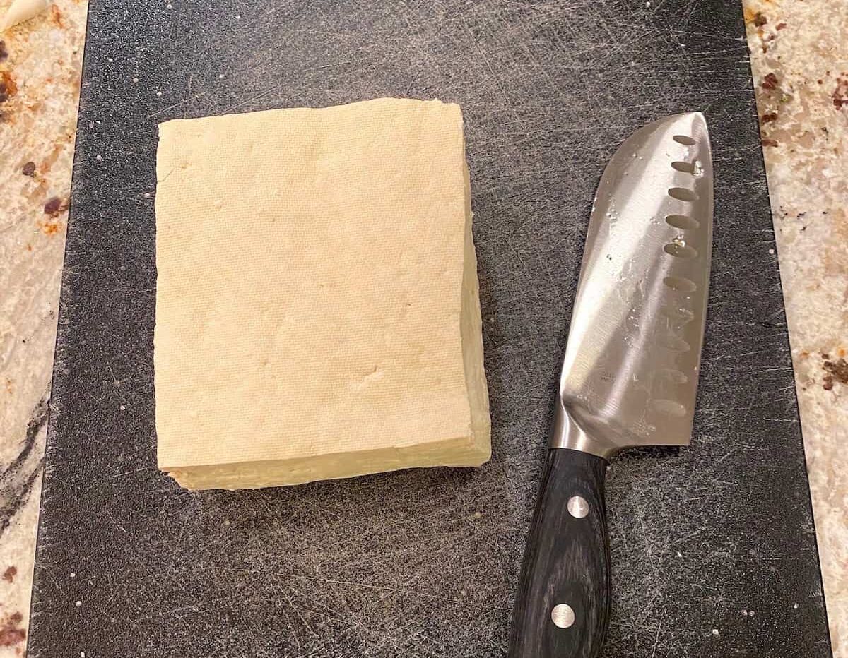 A slab of tofu on a cutting board with a knife on the side.