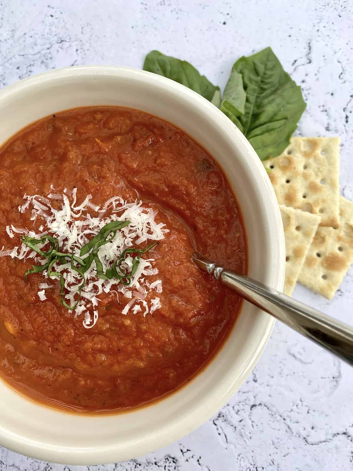 A bowl a tomato basil soup with crackers and fresh basil on the side. 