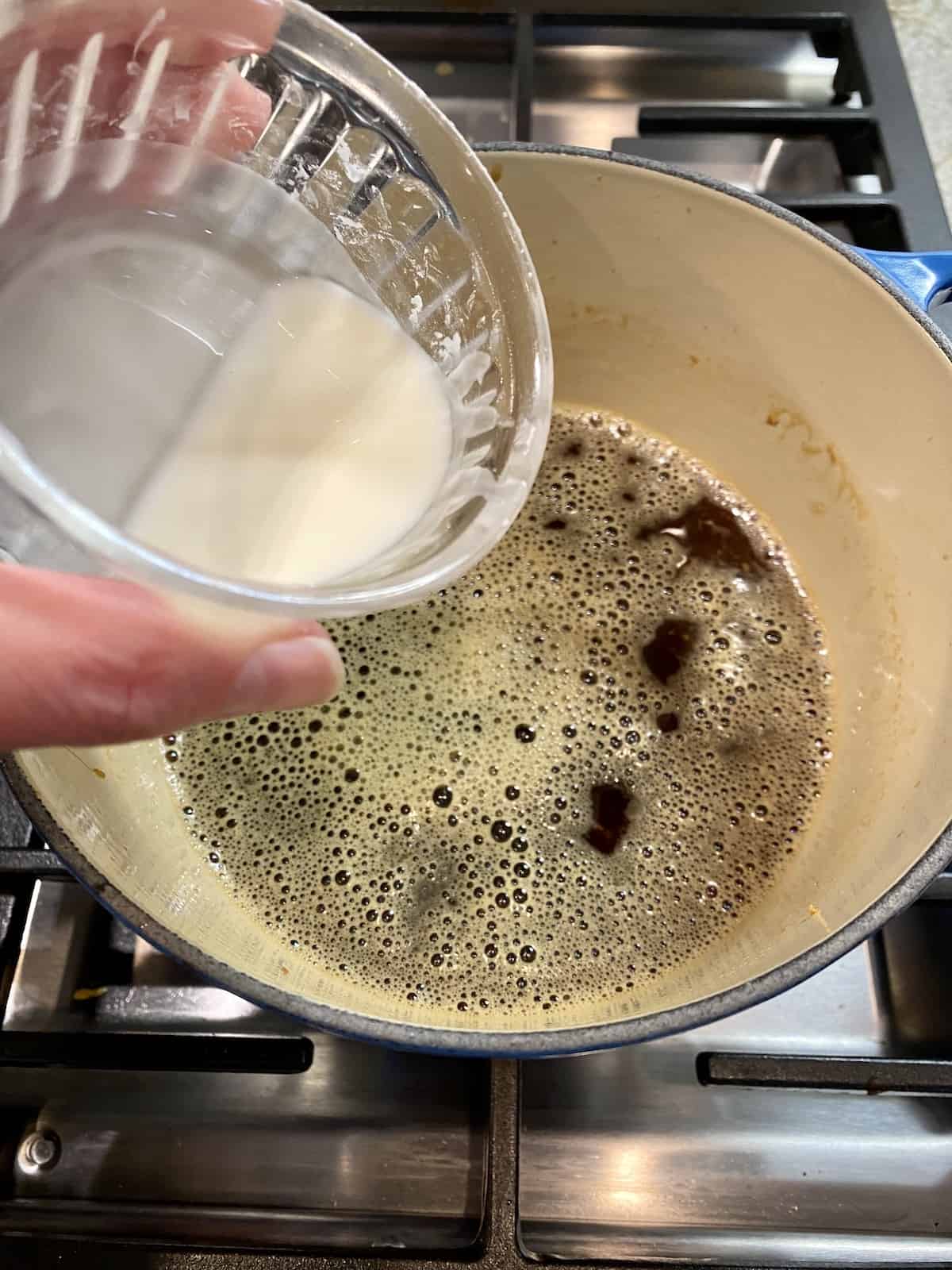 A small container of cornstarch mixed with water, being held over a saucepan with orange sauce on a stove top.