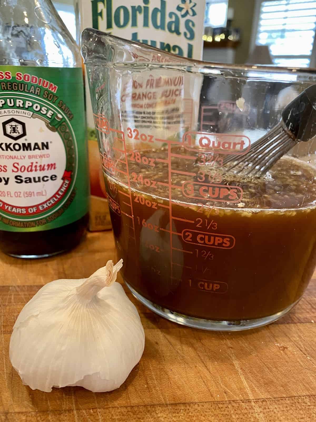 Orange sauce ingredients in a measuring cup, on a wooden cutting board with a bulb of garlic, soy sauce and orange juice.,