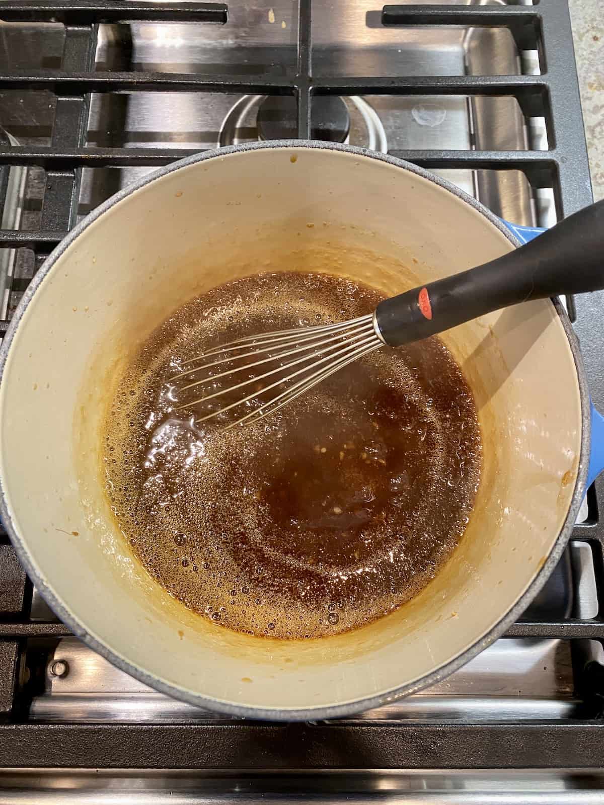 Orange sauce in a saucepan on the stove with a metal whisk on the side.