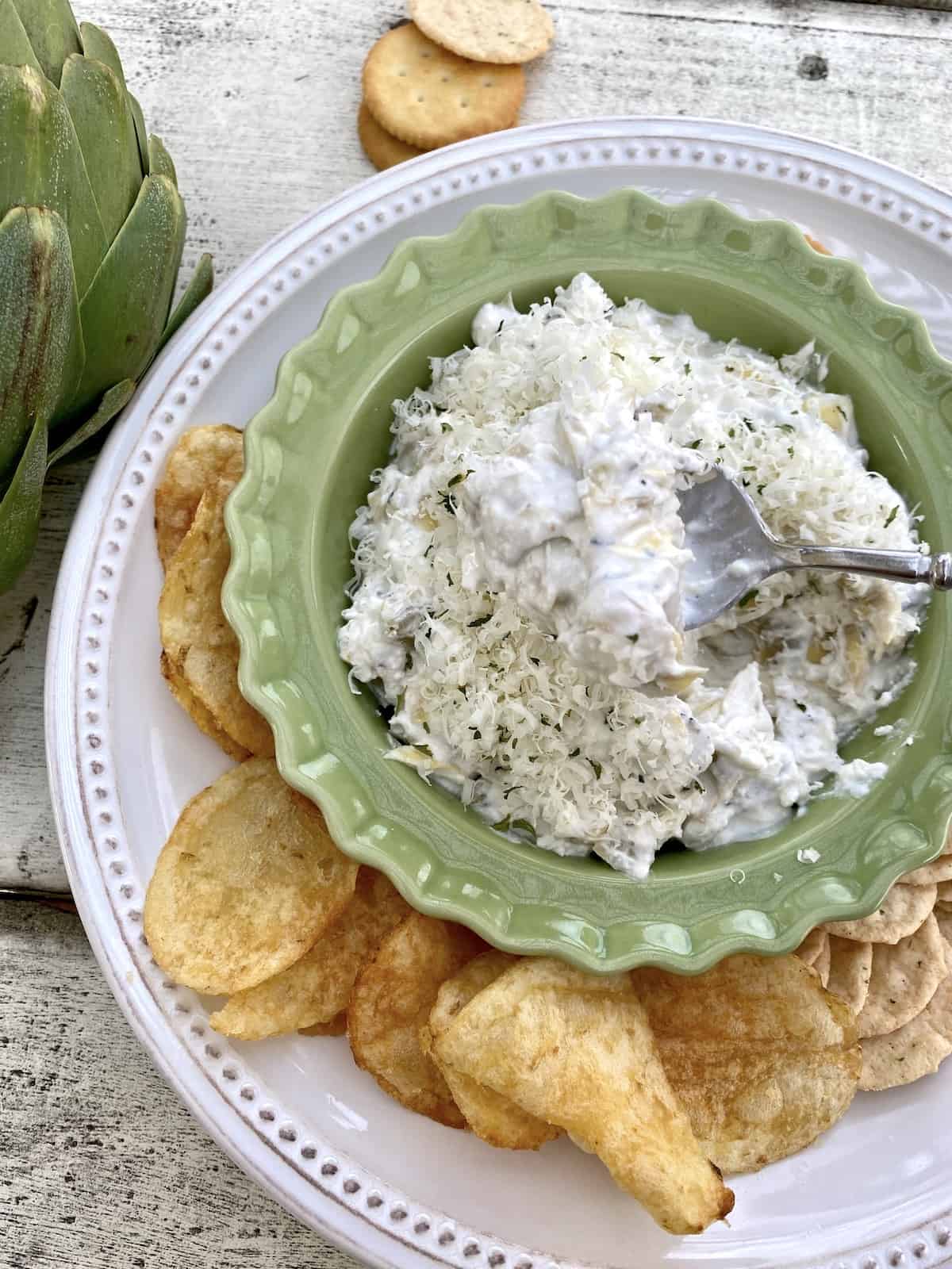 A large plate with chips and crackers and a small bowl of artichoke dip in the center.