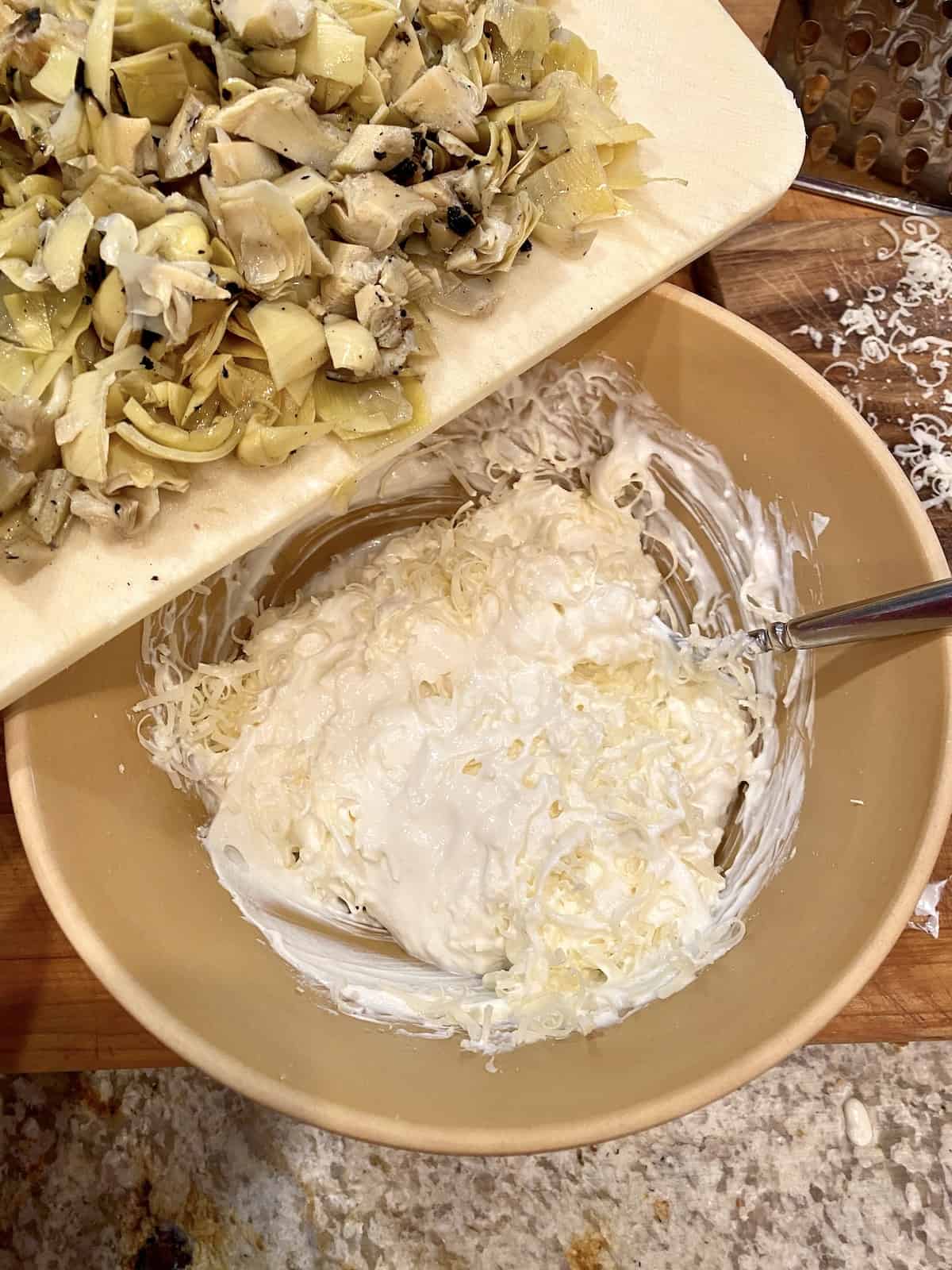 A bowl of creamy ingredients for artichoke dip, with a cutting board of chopped artichoke hearts to the side. 