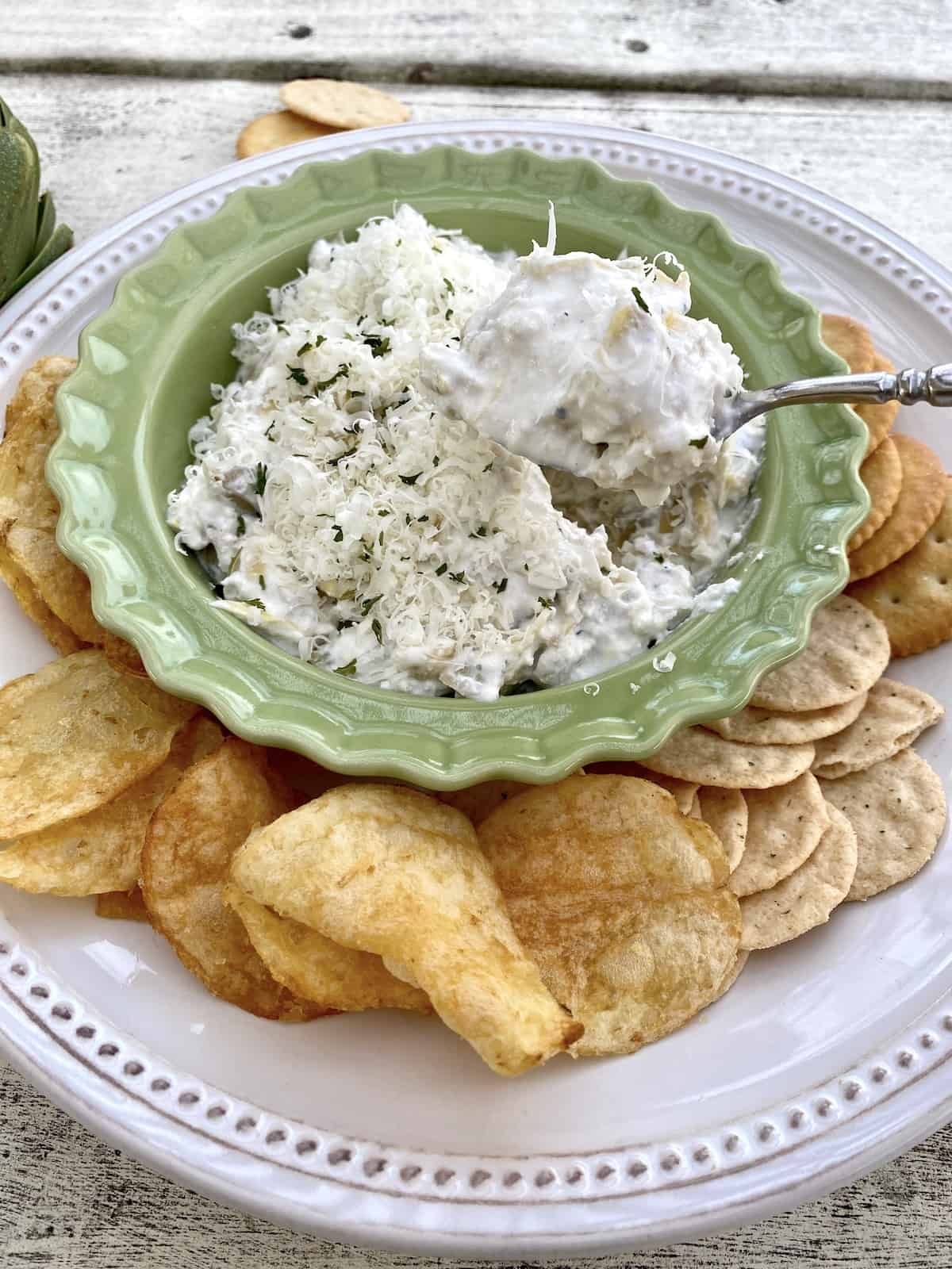 A bowl of dip with a spoon and crackers arranged around the bowl. 