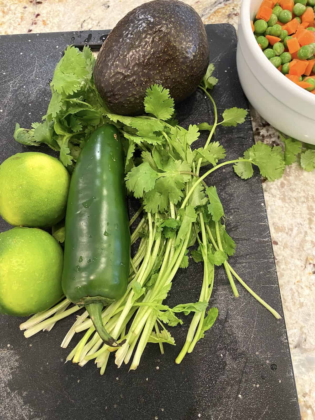 A jalapeno pepper with fresh limes and cilantro on a cutting board next to a bowl of peas and carrots.