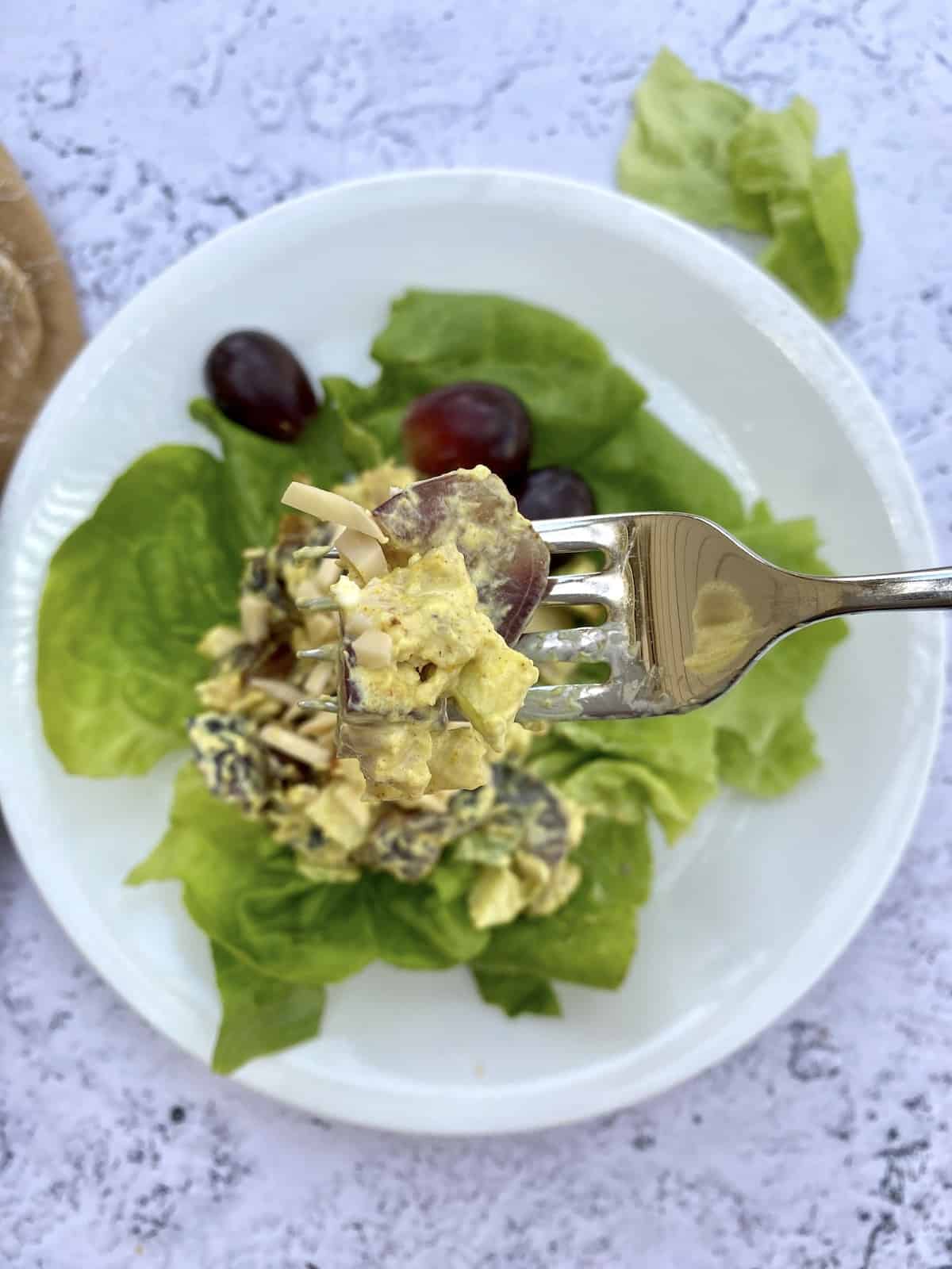 A fork held over a plate of salad with a scoop of chicken curry and grapes.