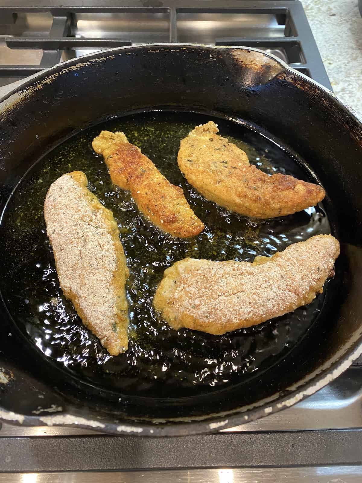 Four chicken tenders in a cast iron skillet, cooking in a small layer of hot oil.