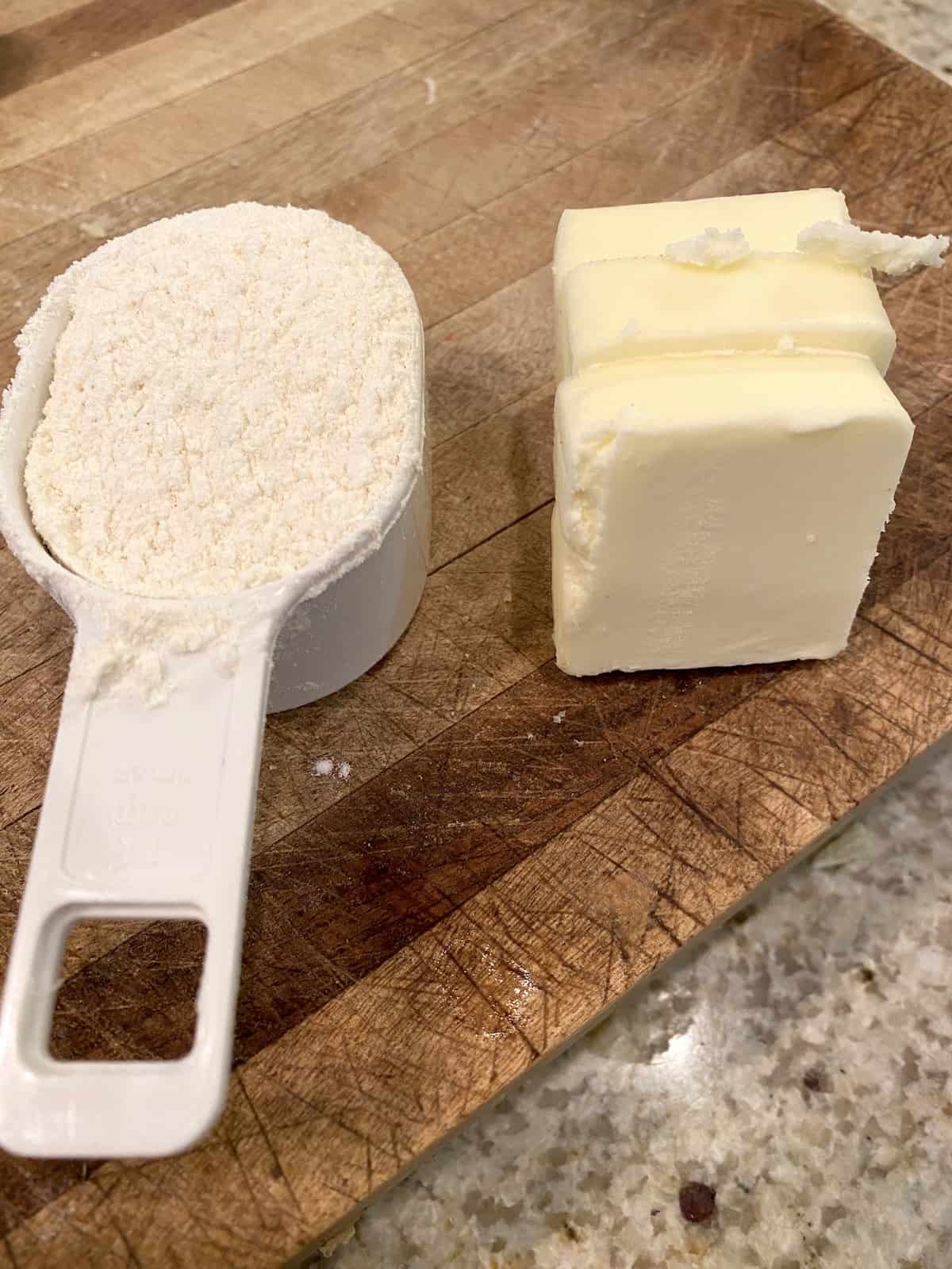 A measuring scoop full of flour and a wedge of butter on a cutting board.