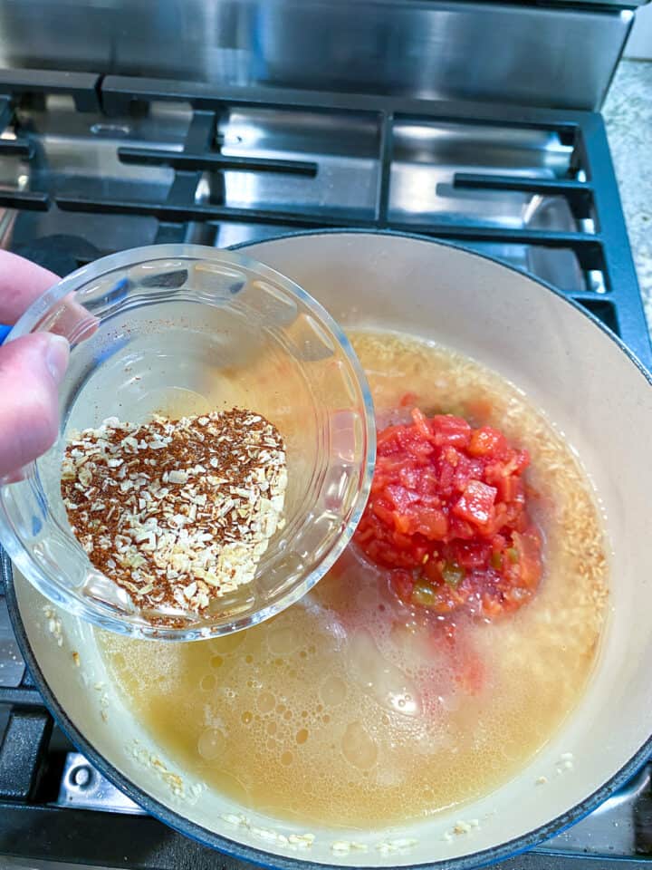 A small dish of spices held over a pan with broth, canned tomatoes and rice.