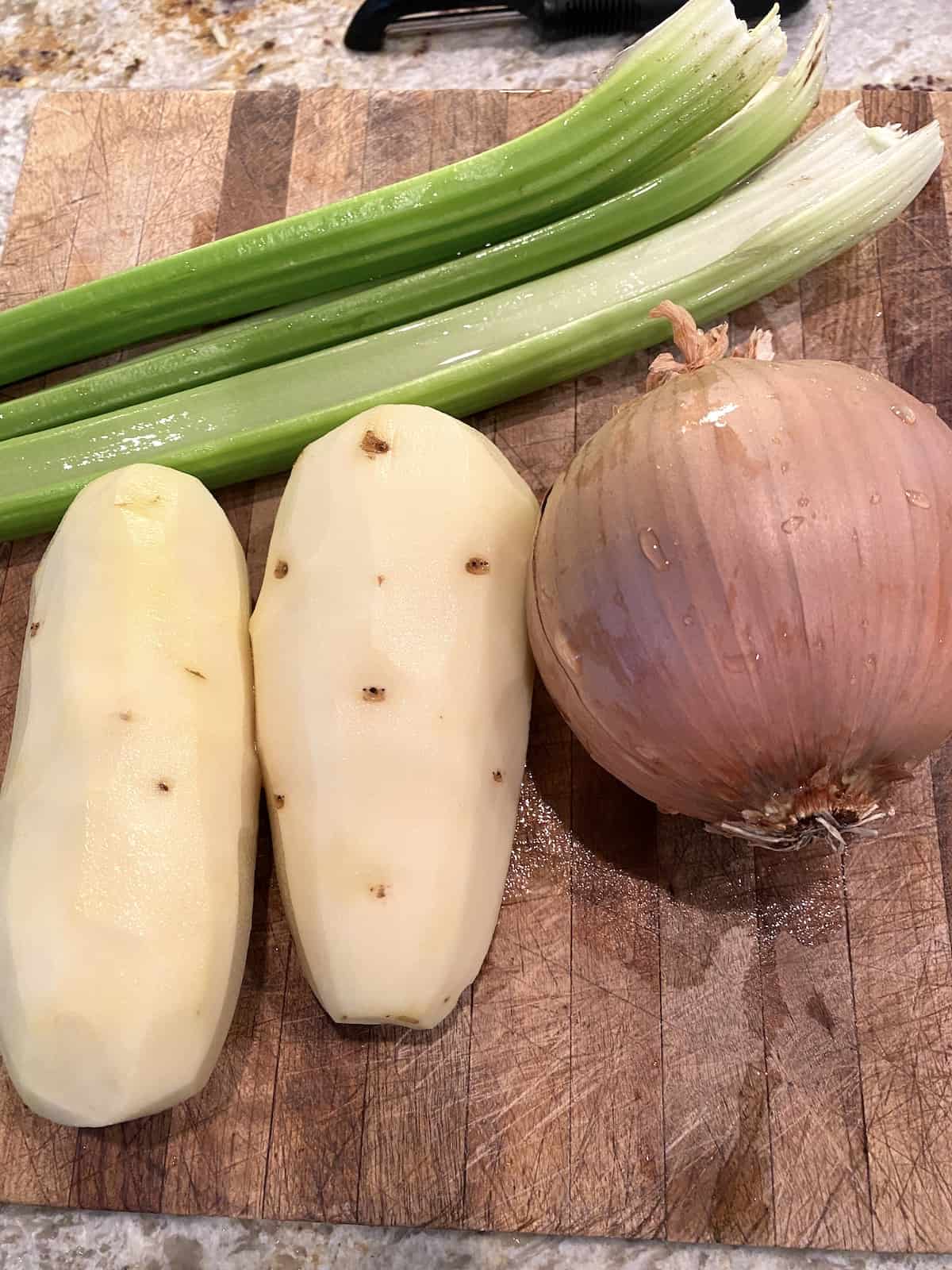 Peeled potatoes, a few stalks of celery and onion on a cutting board.