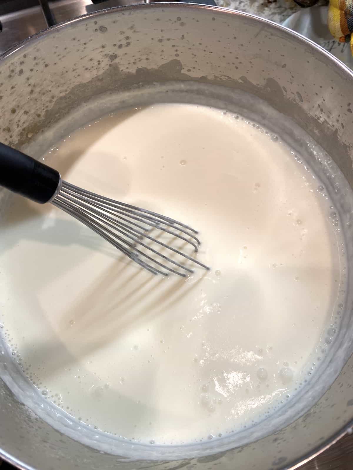 White sauce in a saucepan on the stove with a whisk on the side.