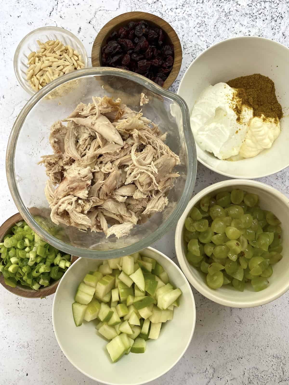 Chicken curry salad ingredients displayed in small bowls. Ingredients include diced apples, grapes, green onions, chicken, dried cranberries and almonds.