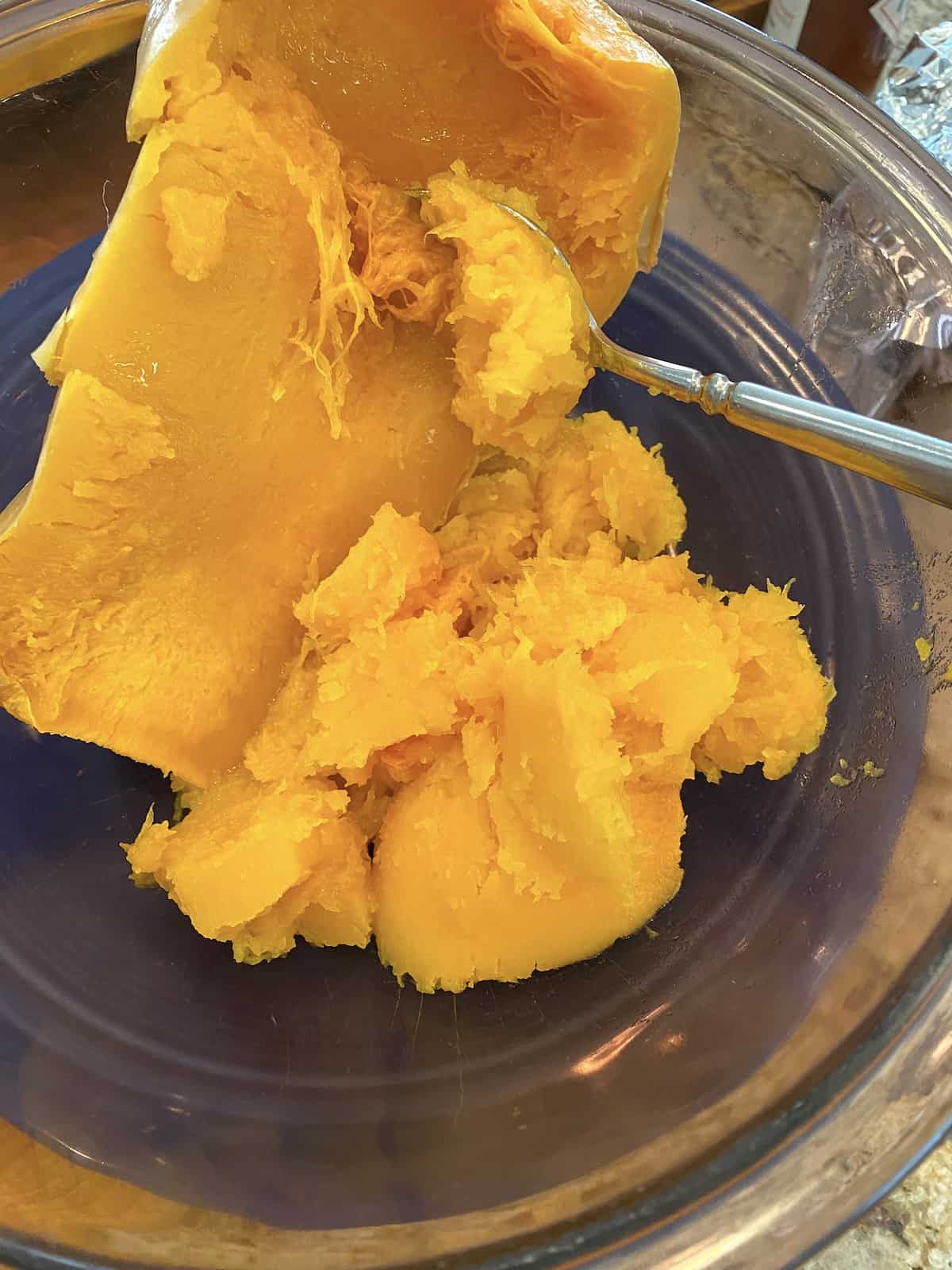 A spoon is scooping out the cooked butternut squash from the peel and into a bowl.