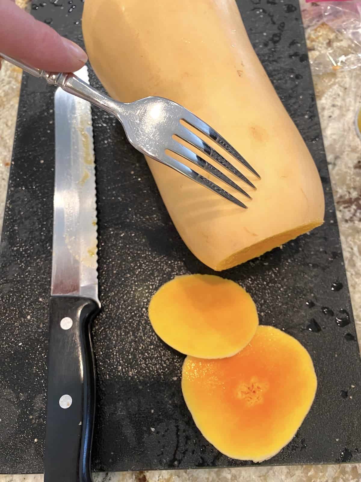 A fork making small holes in the peel of an uncooked butternut squash. The ends are sliced off and by the side as is a knife.