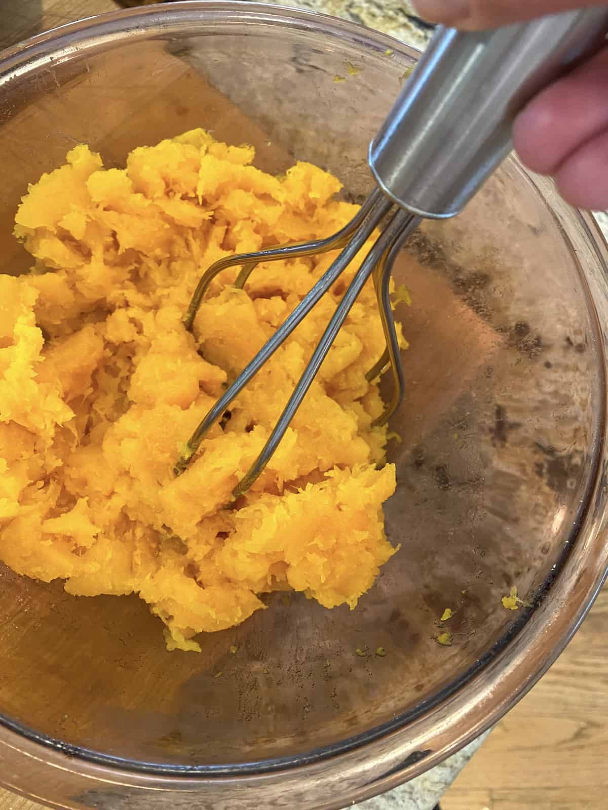 A potato masher being used to mash cooked butternut squash in a medium-sized bowl.