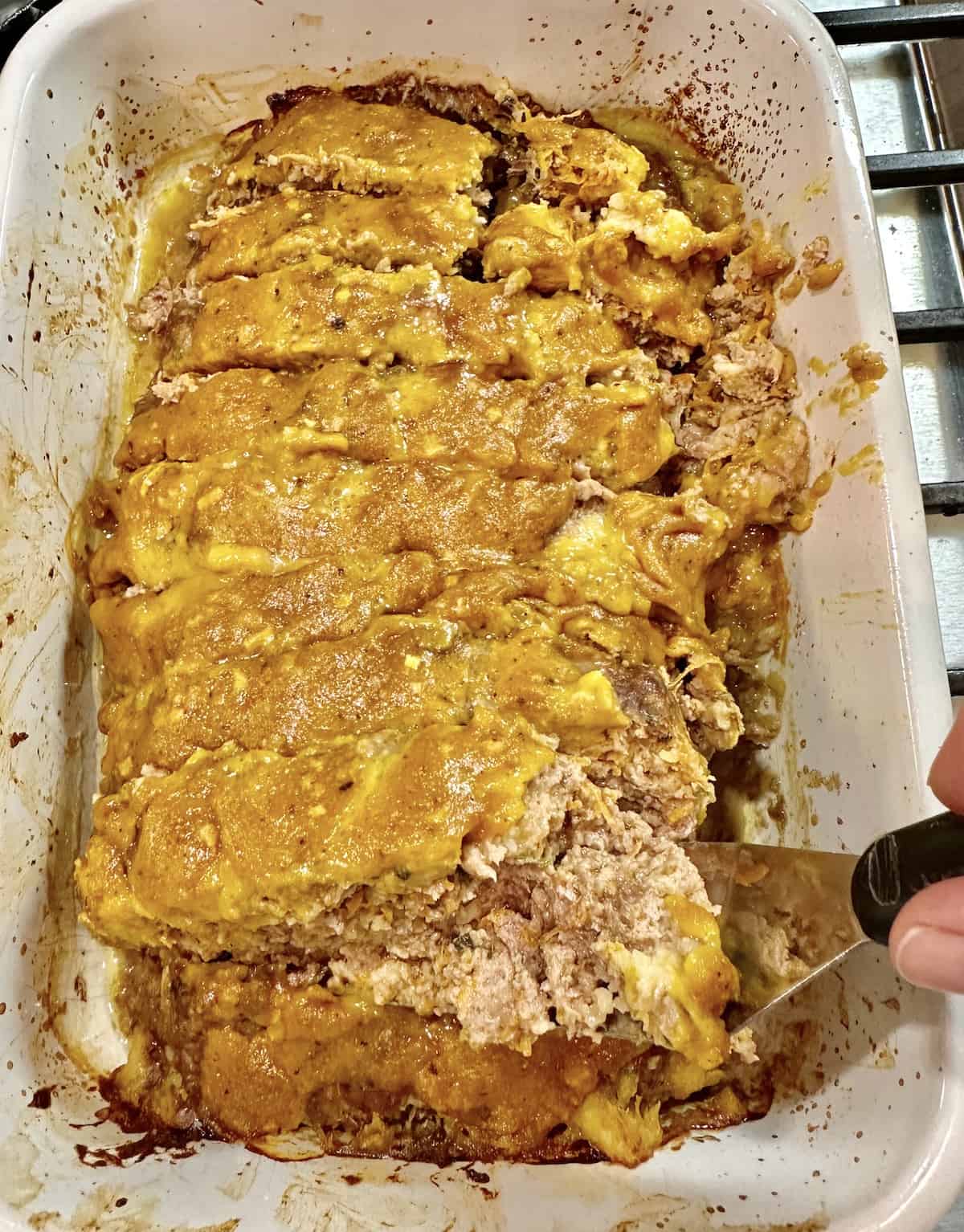 A baked meatloaf with mustard glaze on top and a spatula scooping out a serving of meatloaf.