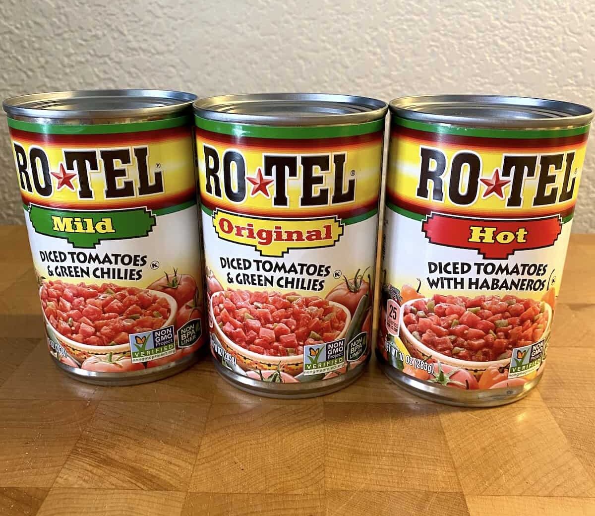 Three cans of Rotel lined up on a cutting board. Each a different variety for spiciness.