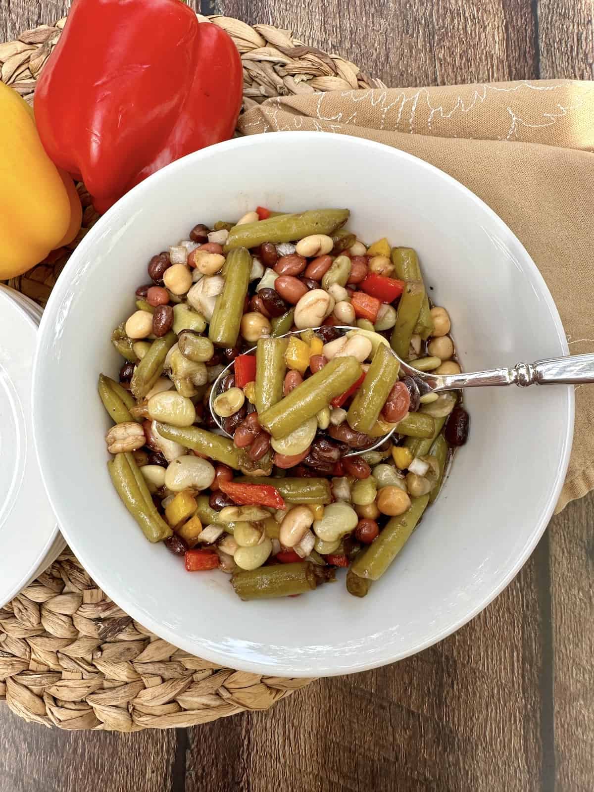 A serving bowl of cold green bean salad with a spoon, on a place mat with two peppers off to the side.