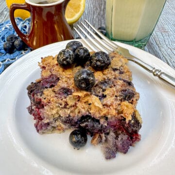 A plate with a fork on the side and a serving of blueberry baked oatmeal in the center with a glass of milk and a small pitcher on the side.