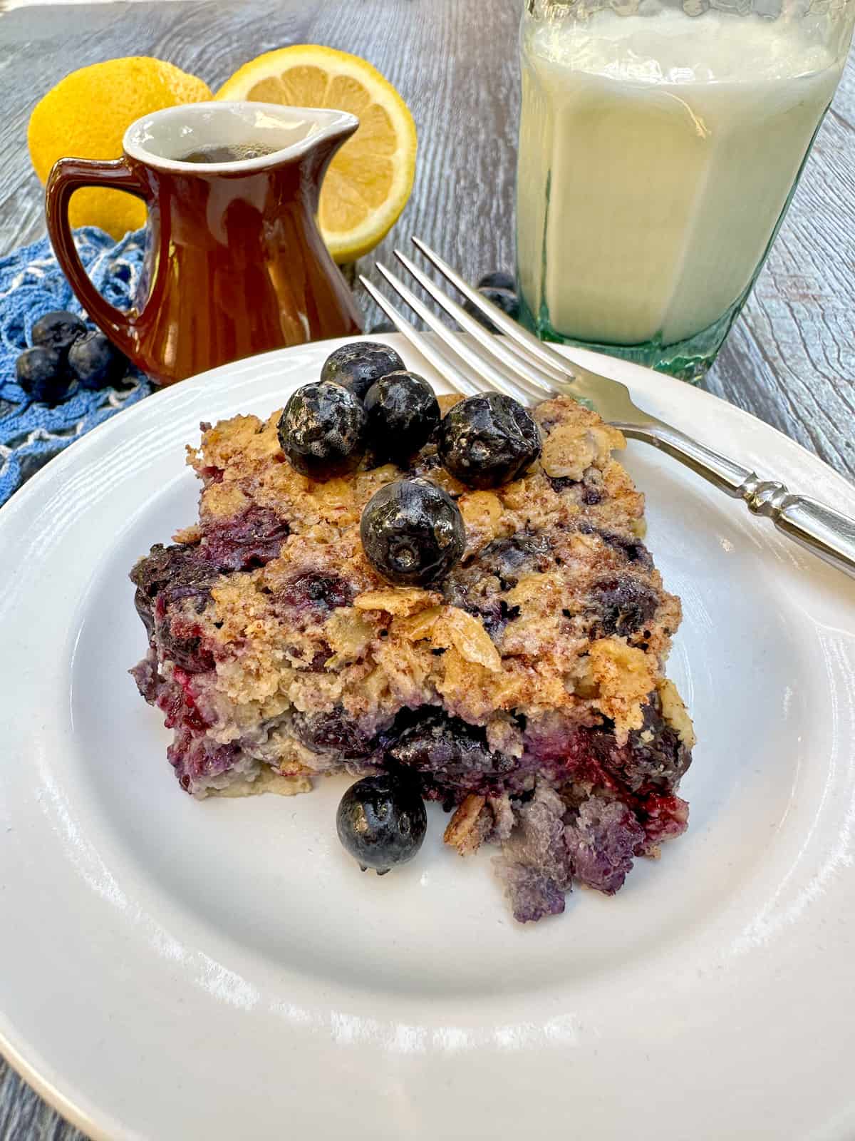 A plate with a fork on the side and a serving of blueberry baked oatmeal in the center with a glass of milk and a small pitcher on the side.