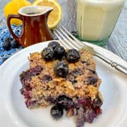 A plate with a portion of blueberry oatmeal in the center with milk and syrup in a glass and small pitcher on the side.
