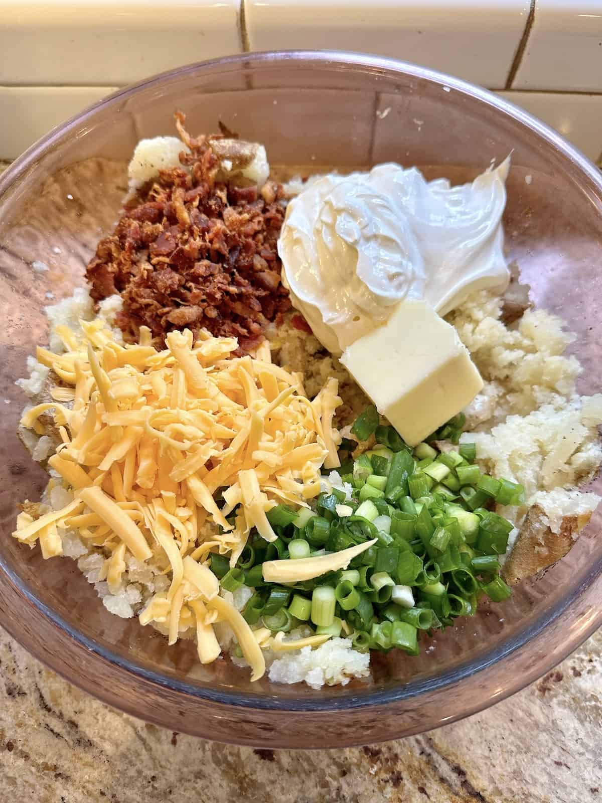 Cheese, green onions, cooked bacon, sour cream and butter arranged on top of mashed, baked potatoes in a bowl.