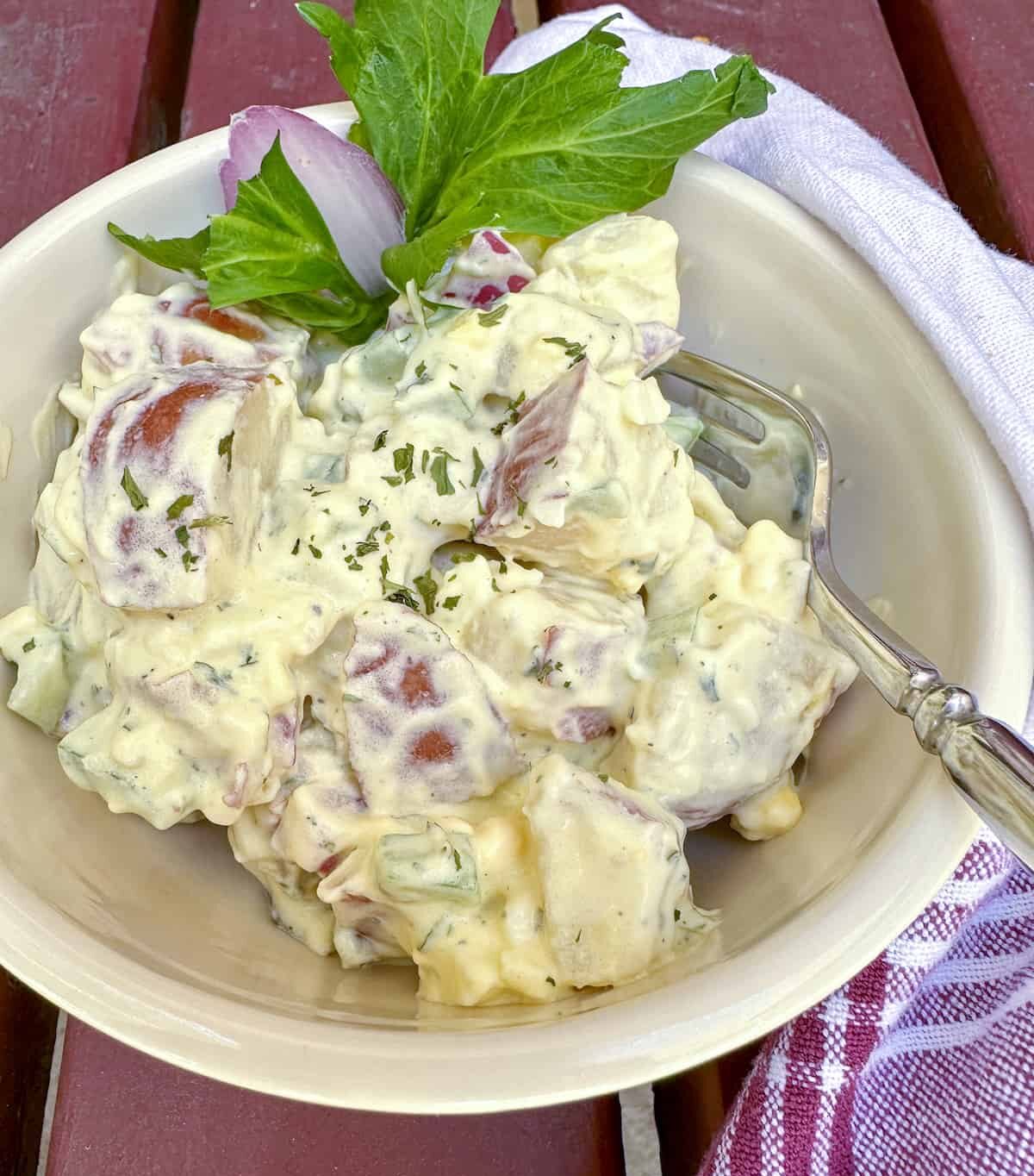 Red potato salad in a bowl with a sprig of celery, a fork in the bowl and a napkin on the side.