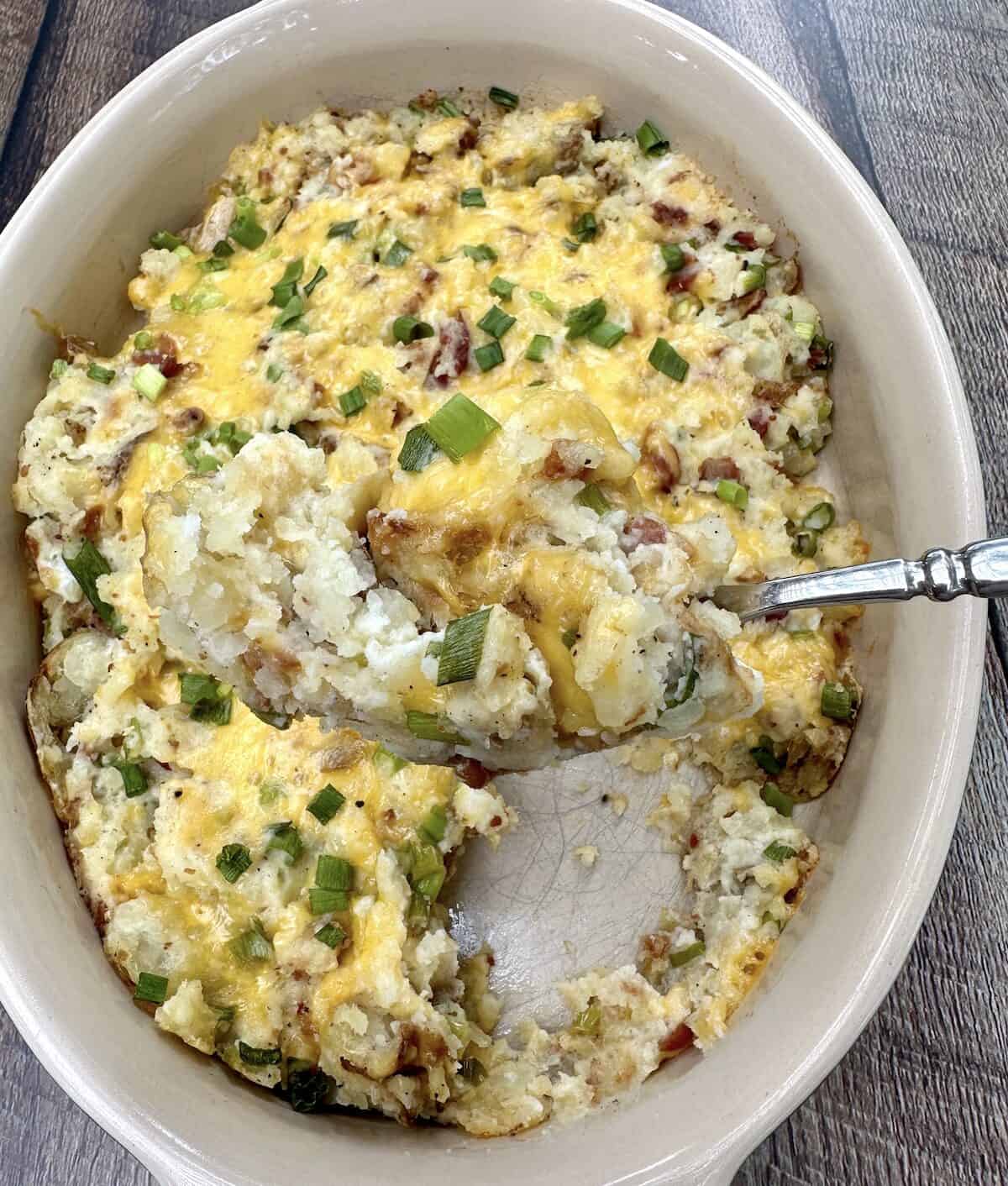 A large baking dish filled with twice baked mashed potatoes and a spoon scooping up a serving.