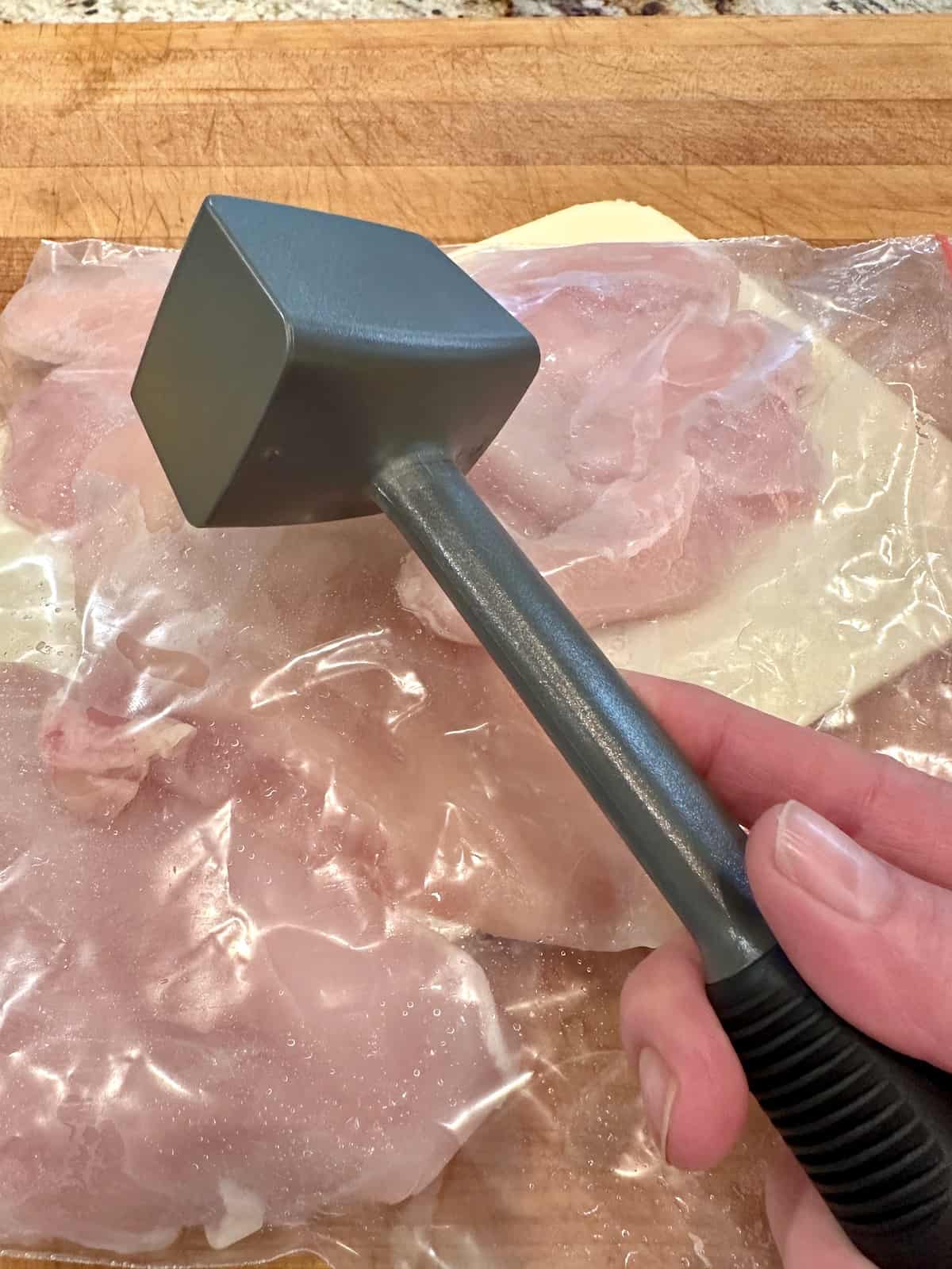 A meat mallet held over a plastic bag of sliced chicken.