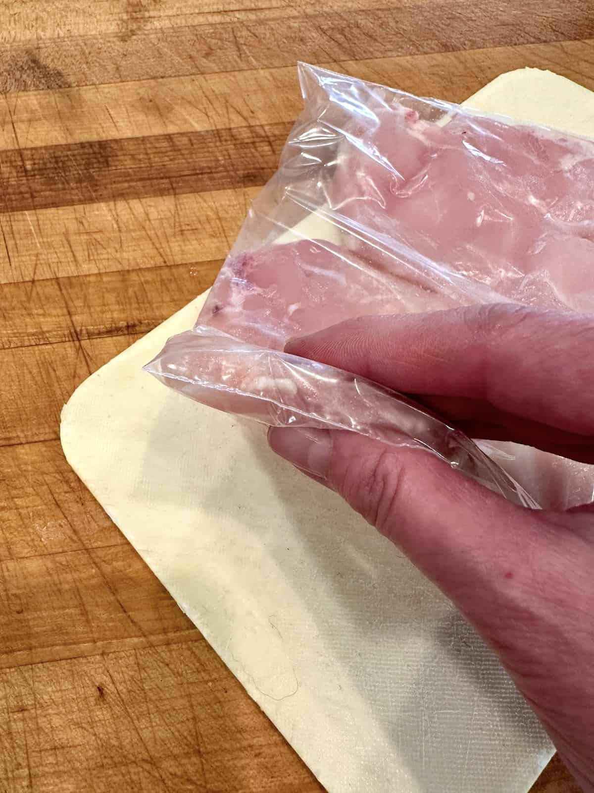 A thinly sliced and tenderized chicken breast in a plastic bag, held by a hand.