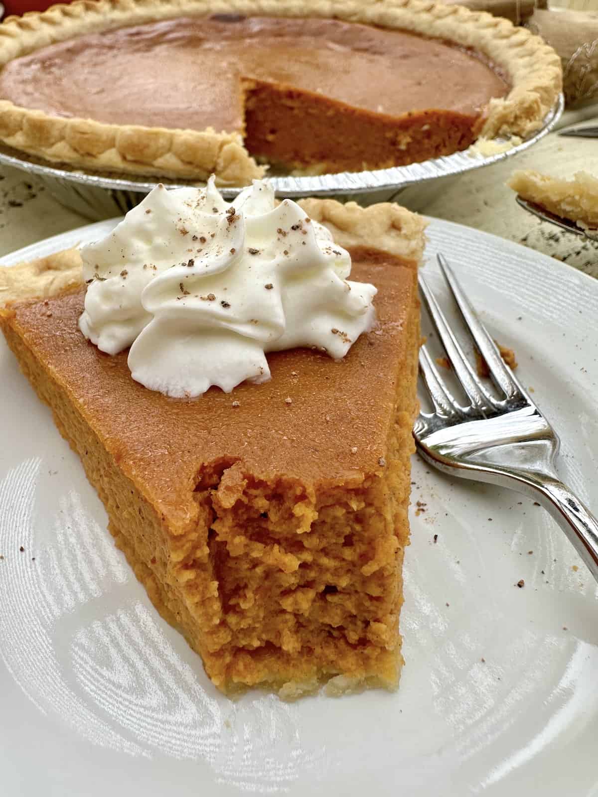 A slice of pumpkin pie with the pie in the background.