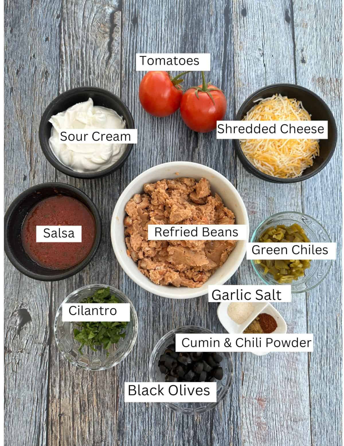 Labeled ingredients for five-layer taco dip on a wooden table.