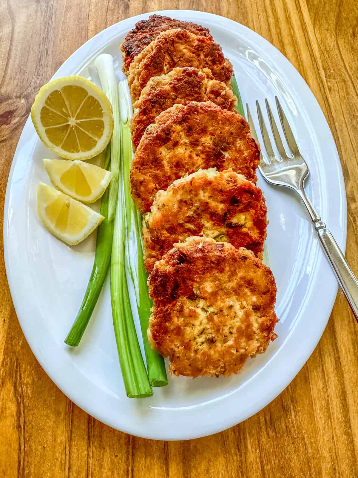 Cooked salmon patties on a plate with scallions, lemons and a fork.