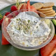 Garlic and herb dip in a dish on a plate with chopped vegetables and crackers.