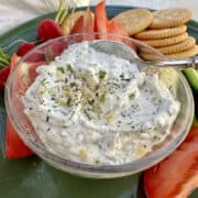 Garlic and herb dip in a dish on a plate with chopped vegetables and crackers.