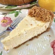 A slice of vanilla cheesecake on a plate with the rest of the cake and a lemon in the background.