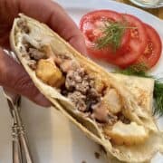 One half of a French taco held by a hand over a plate with silverware, sliced tomato and dill.