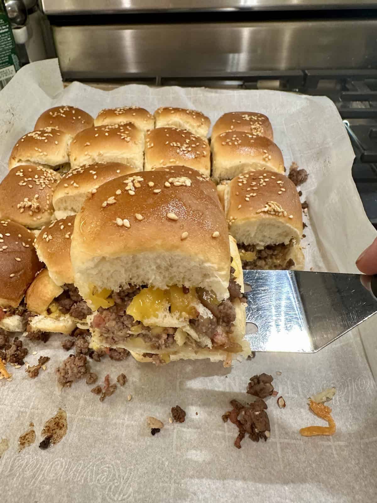 A spatula serving a cheeseburger slider from a baking pan on the stovetop.
