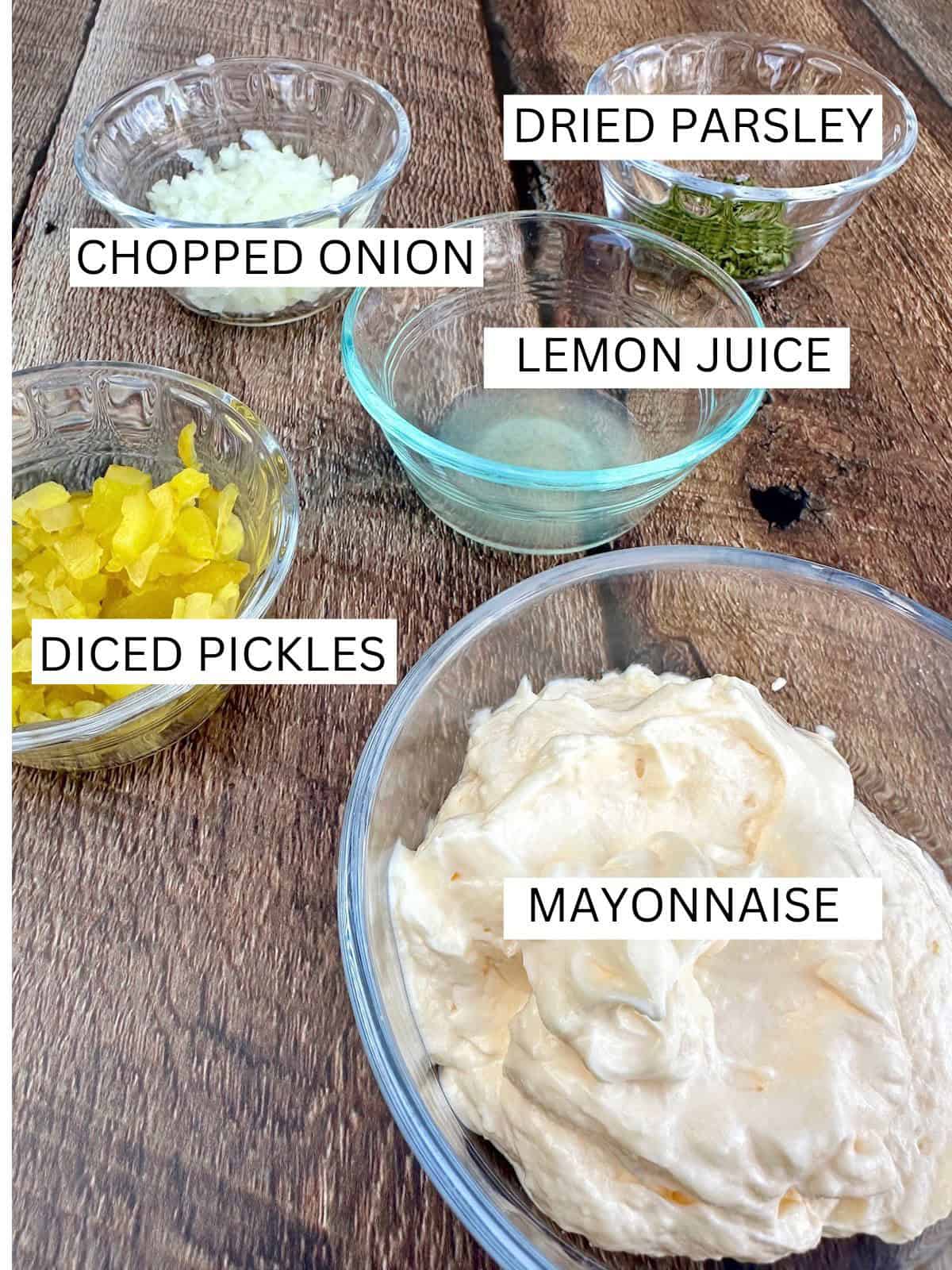 Tartar sauce ingredients in small bowls on a wooden board.
