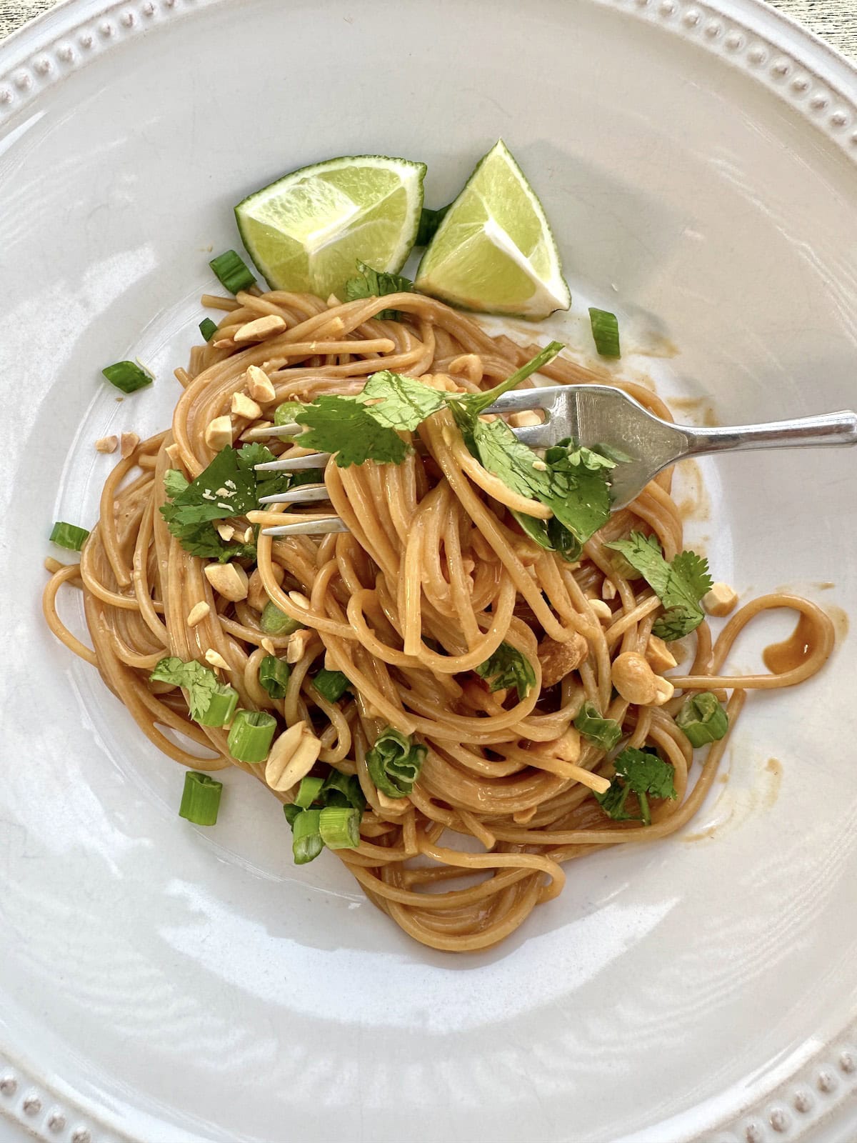 Spicy peanut noodles on a serving plate with lime, peanuts, onion and cilantro.