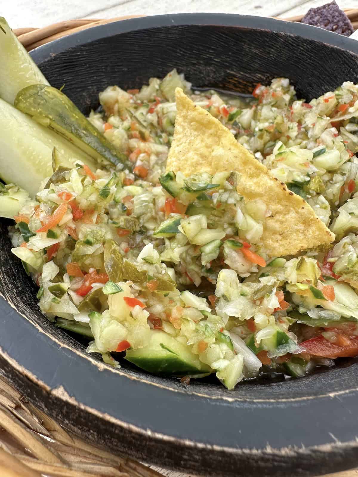 A serving bowl of pickle de gallo with pickles and a tortilla chip on top,.