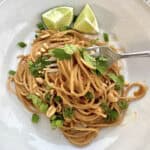 A serving of spicy peanut noodles on a plate with peanuts, onion and cilantro.