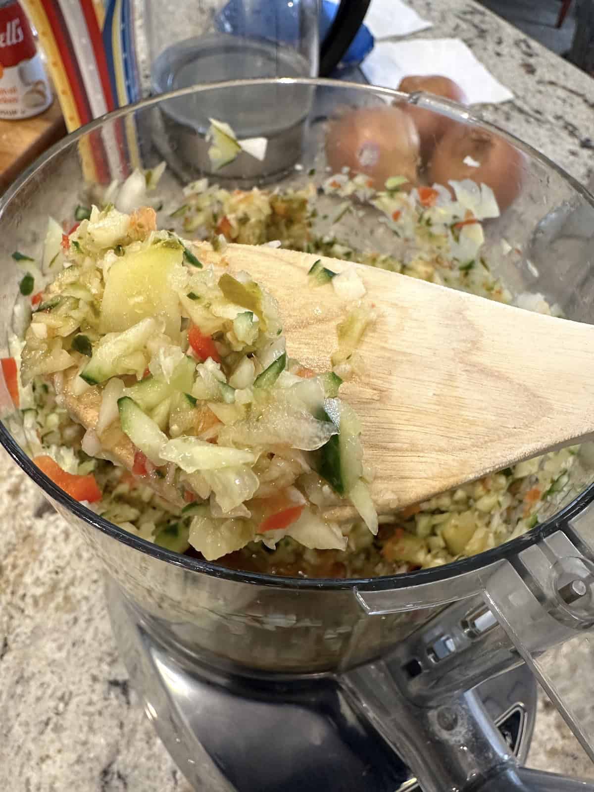 A wooden spoon with a scoop of chopped pickles and cucumbers in a food processor.