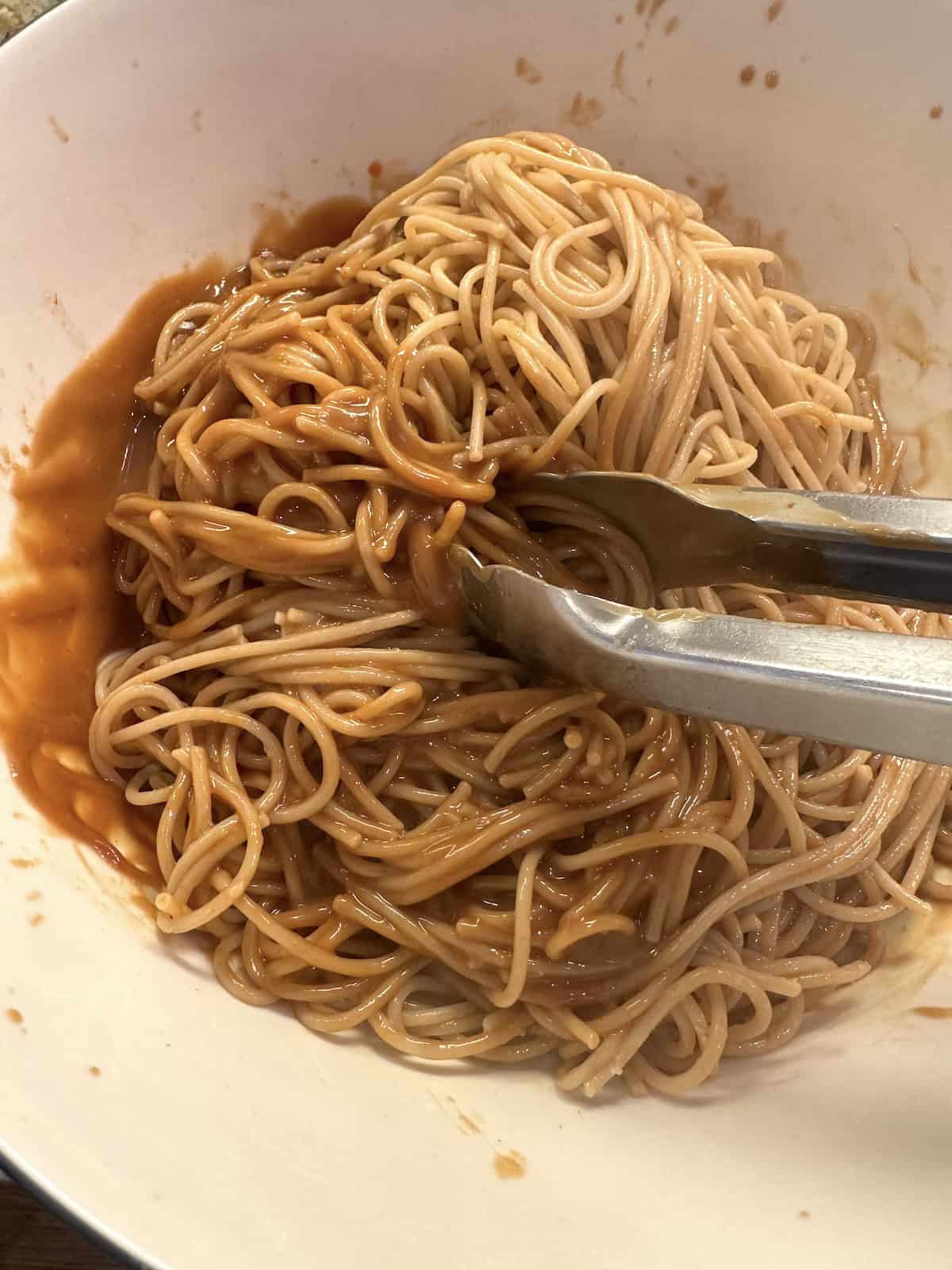 Cooked spaghetti noodles in a bowl with sauce and tongs.