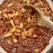 Franks, baked beans and pineapple slices in a baking dish with a wooden spoon.