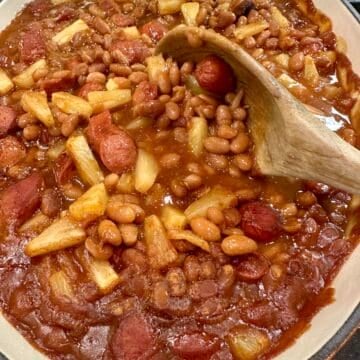 Franks, baked beans and pineapple slices in a baking dish with a wooden spoon.