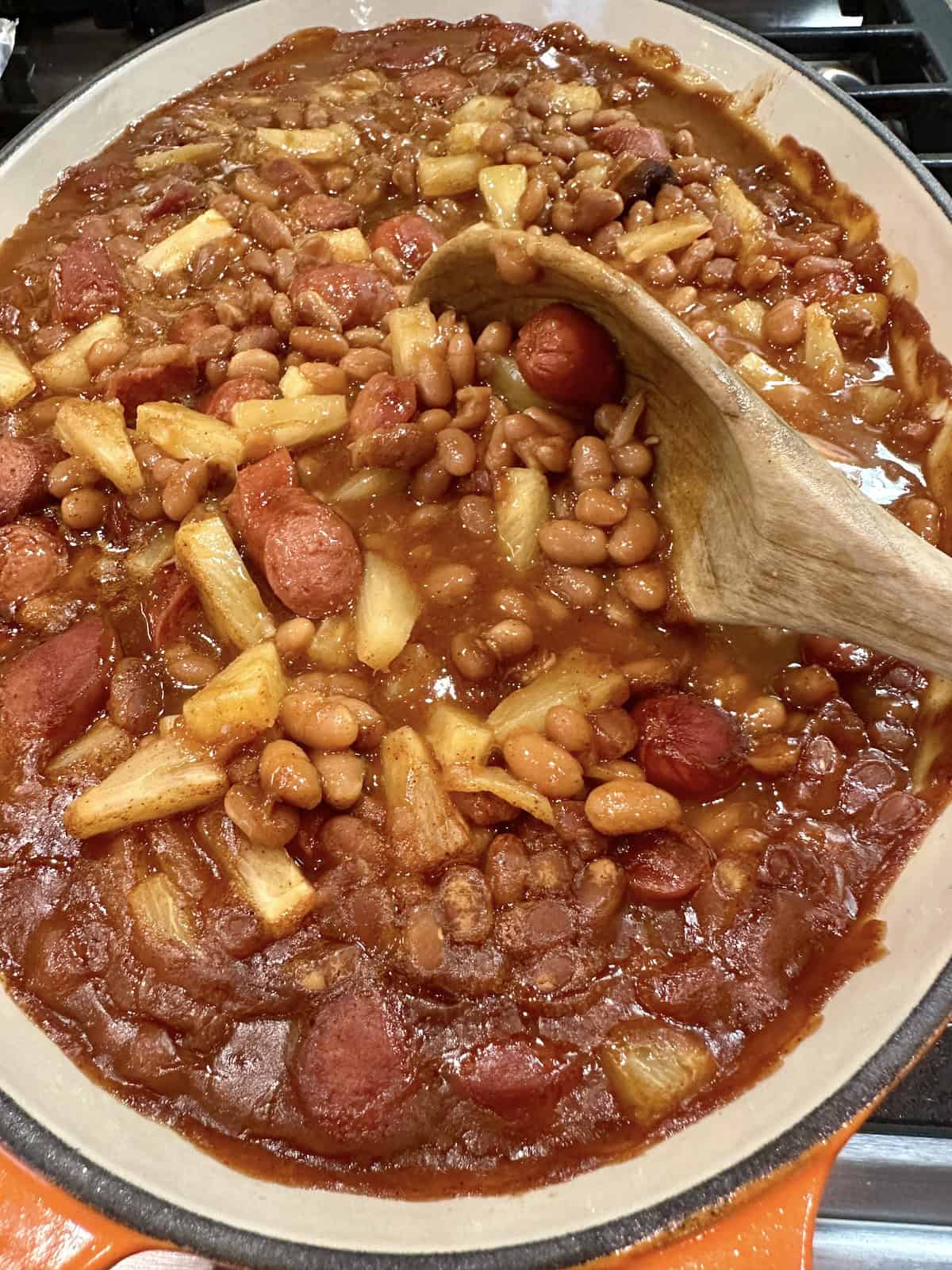 Franks, baked beans and pineapple slices in a baking dish with a wooden spoon.