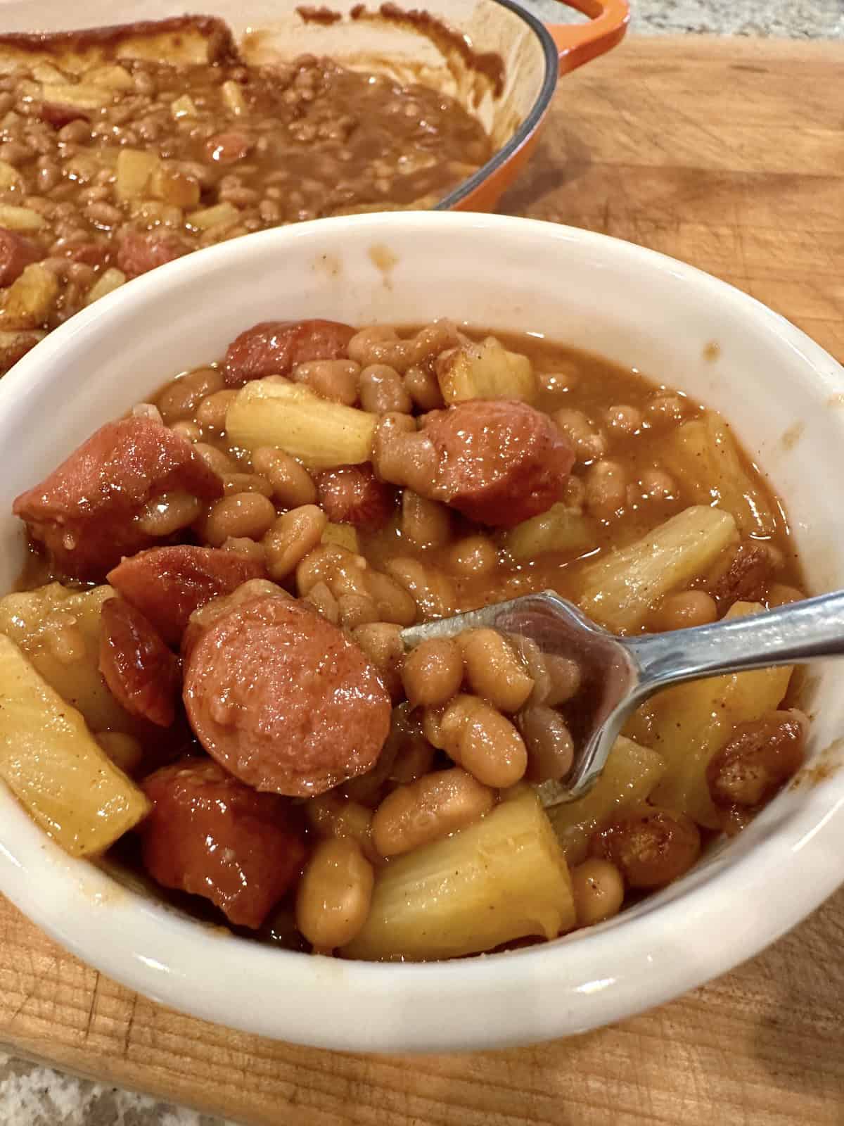 A serving bowl of baked beans, hot dogs and pineapple.