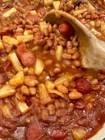 Baked beans, franks and pineapple pieces in a baking dish with a spoon.