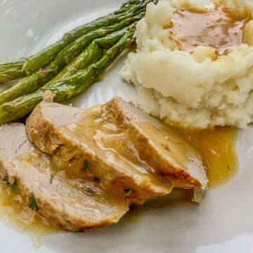 A serving of sliced pork tenderloin with orange sauce on a plate with mashed potatoes and asparagus.